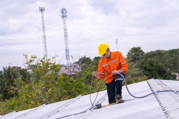 Técnico em um uniforme de segurança laranja e capacete manuseando um cabo elétrico em um telhado. 